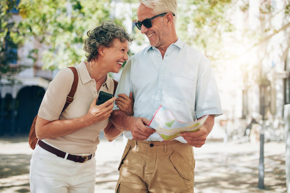 Happy couple, could be 55+ are walking on the street, laughing. She has a phone and he has a map. They could be thinking of retiring to las vegas and doing research of the neighborhoods.