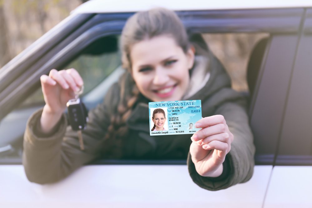 Woman is holding a real estate license and smiling out her car window