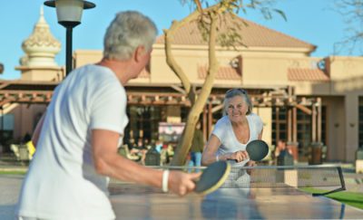 2 grey haired people, a man and a woman, who look to be 55+ are playing ping pong outside with a clubhouse behind them