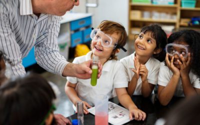 Children at an elementary school are wearing goggles and look up at the teacher smiling as he holds and explains a science project