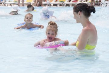 Kids are swimming with tubes and a mom is standing close by holding one of the tubes with a child in it