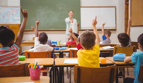 elementary school classroom shows the teacher up front pointing to kids who have their hands raised