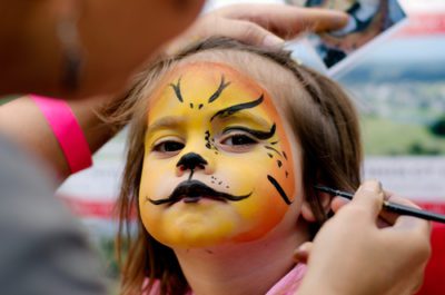 Small child, age 3-5 has her face painted in orange and yelow with whiskers, like they might do at a Springs Preserve Event